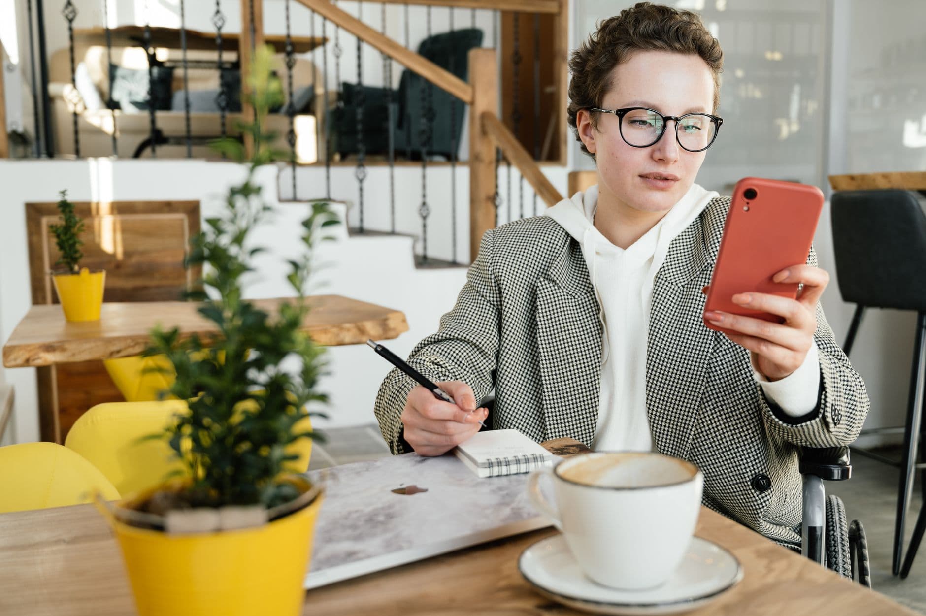 Melhores apps de bloqueio de redes sociais para focar Concentrated businesswoman in formal wear and eyeglasses using wheelchair browsing smartphone and taking notes in copybook while working in cafe