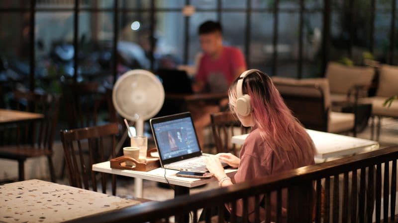Apps de meditação para pausas no trabalho remoto A woman with headphones edits a video on her laptop in a cozy café environment, Vietnam.