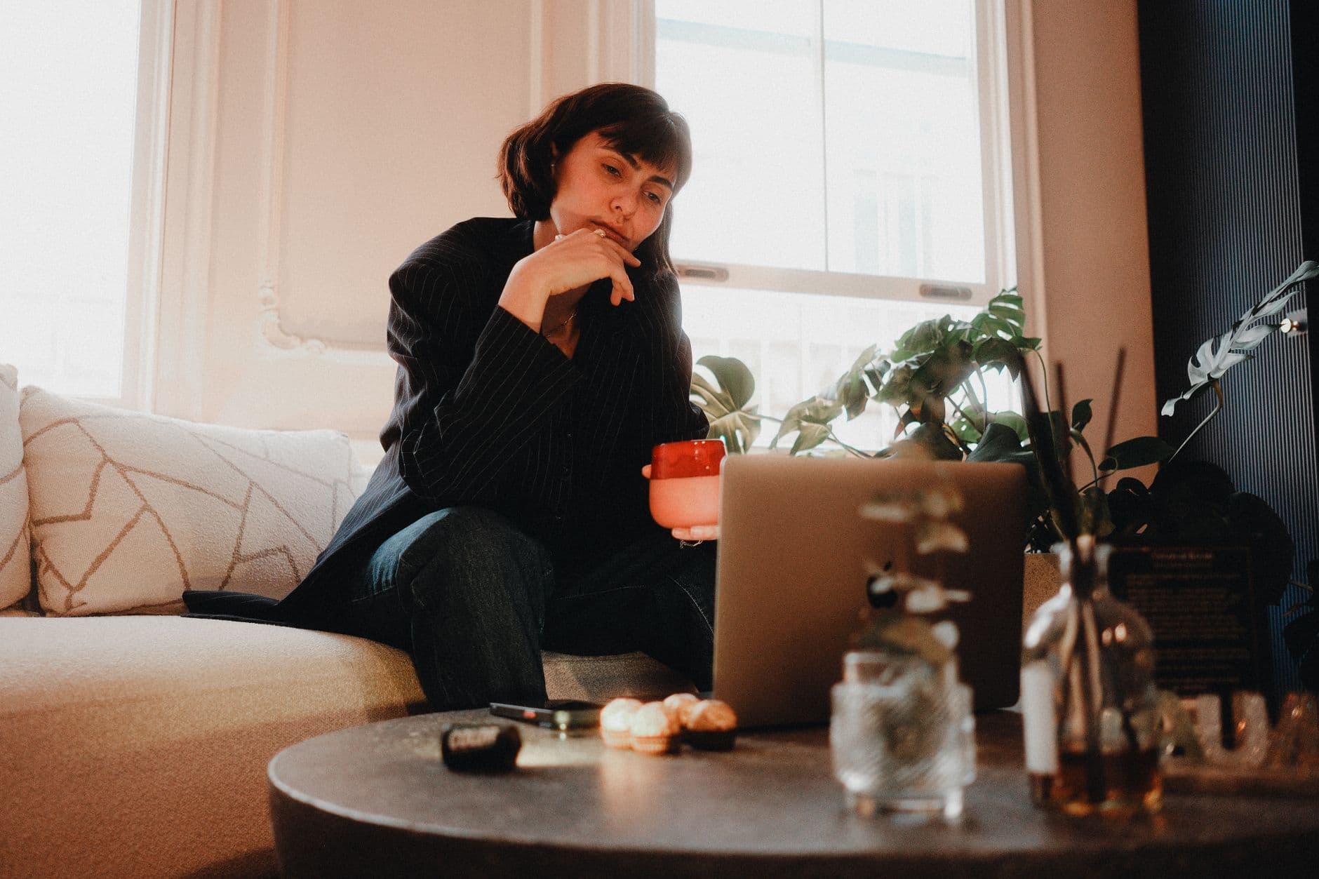Burnout no home office: como prevenir Woman contemplating at home with coffee mug and laptop on table. Cozy indoor setting.