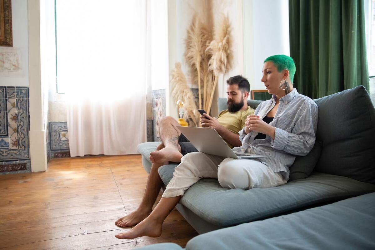 Casal sentado no sofá de casa trabalhando com notebook e celular