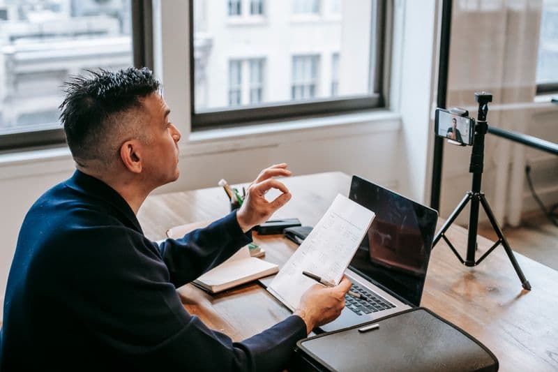 Como aproveitar viagens trabalhando remoto Man using laptop and smartphone for a video call in an office setting.