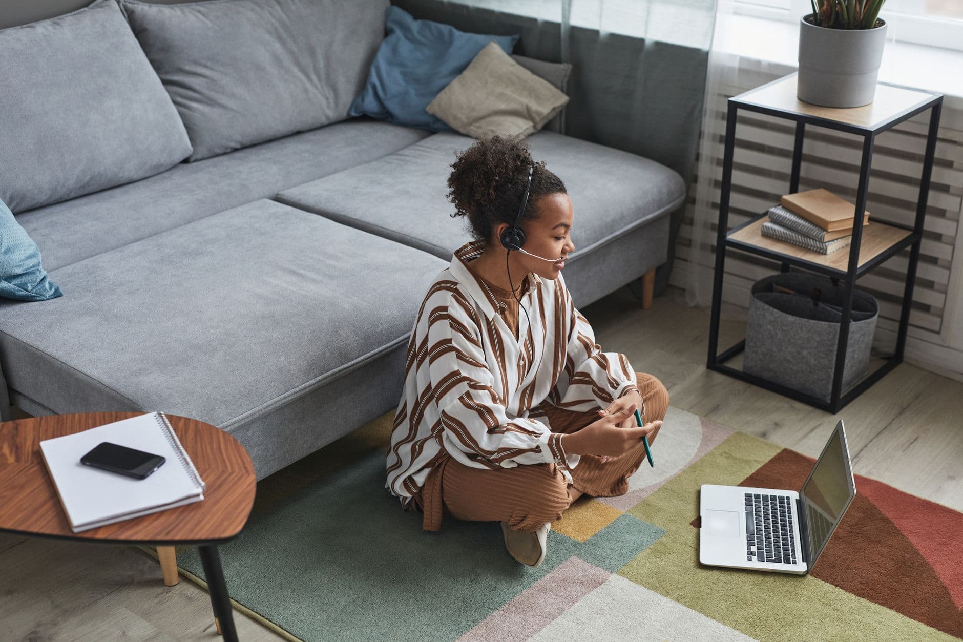 Como lidar com interrupções da família no home office African American woman working remotely at home, sitting on the floor with laptop and headset.