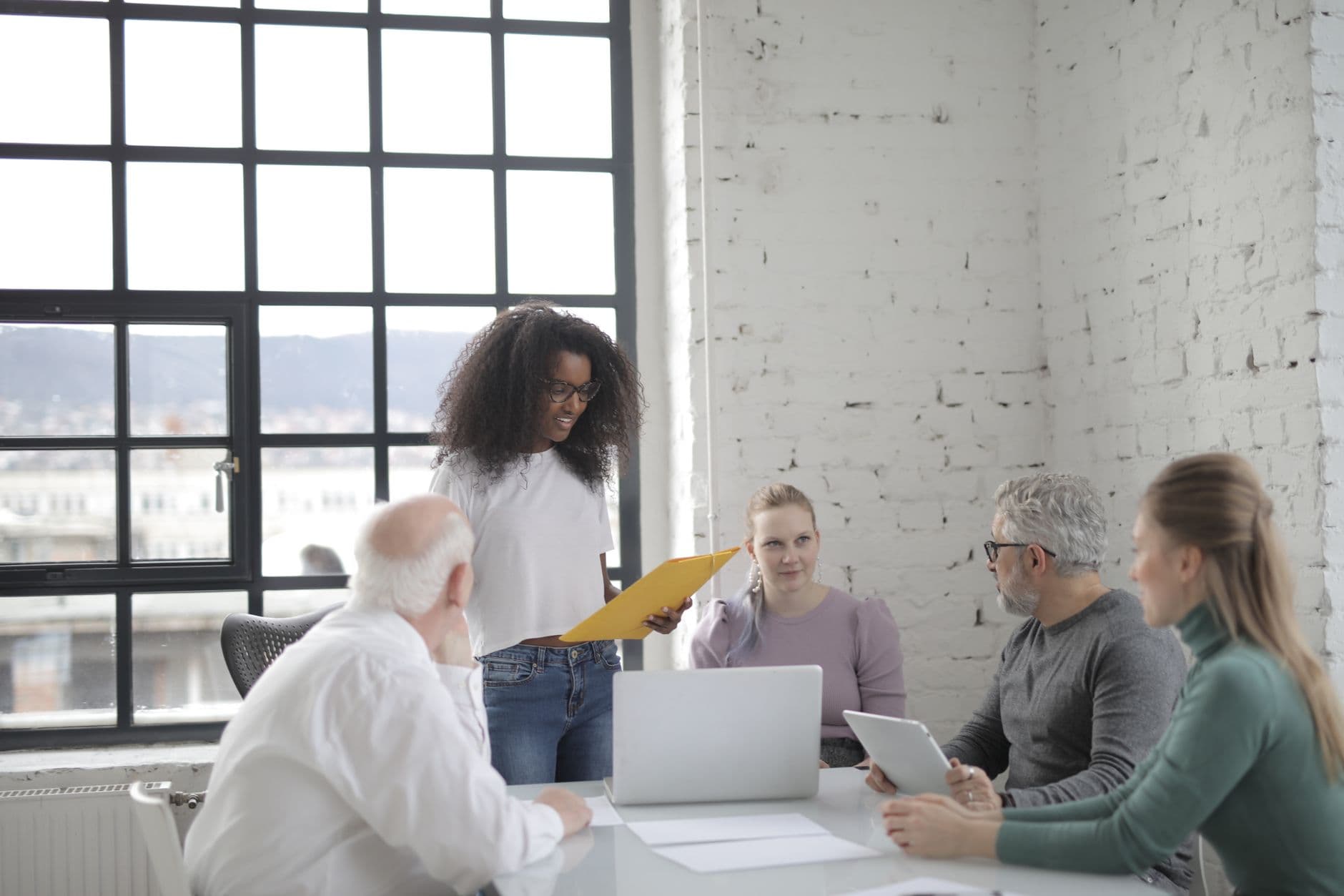 Como organizar arquivos no trabalho remoto Concentrated ethnic woman reading documents while standing near table and explaining details of project to colleagues in modern office near window