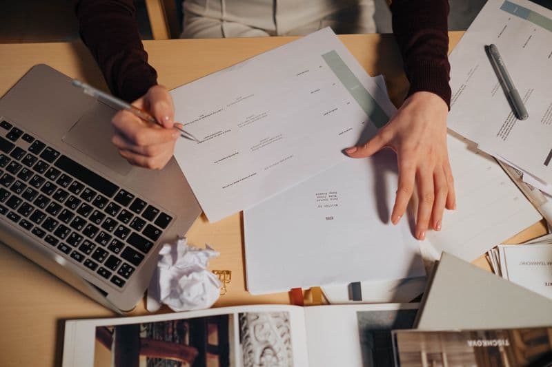 Como pedir aumento de escopo em contrato freelancer A person taking notes and working on a laptop surrounded by documents on a desk.