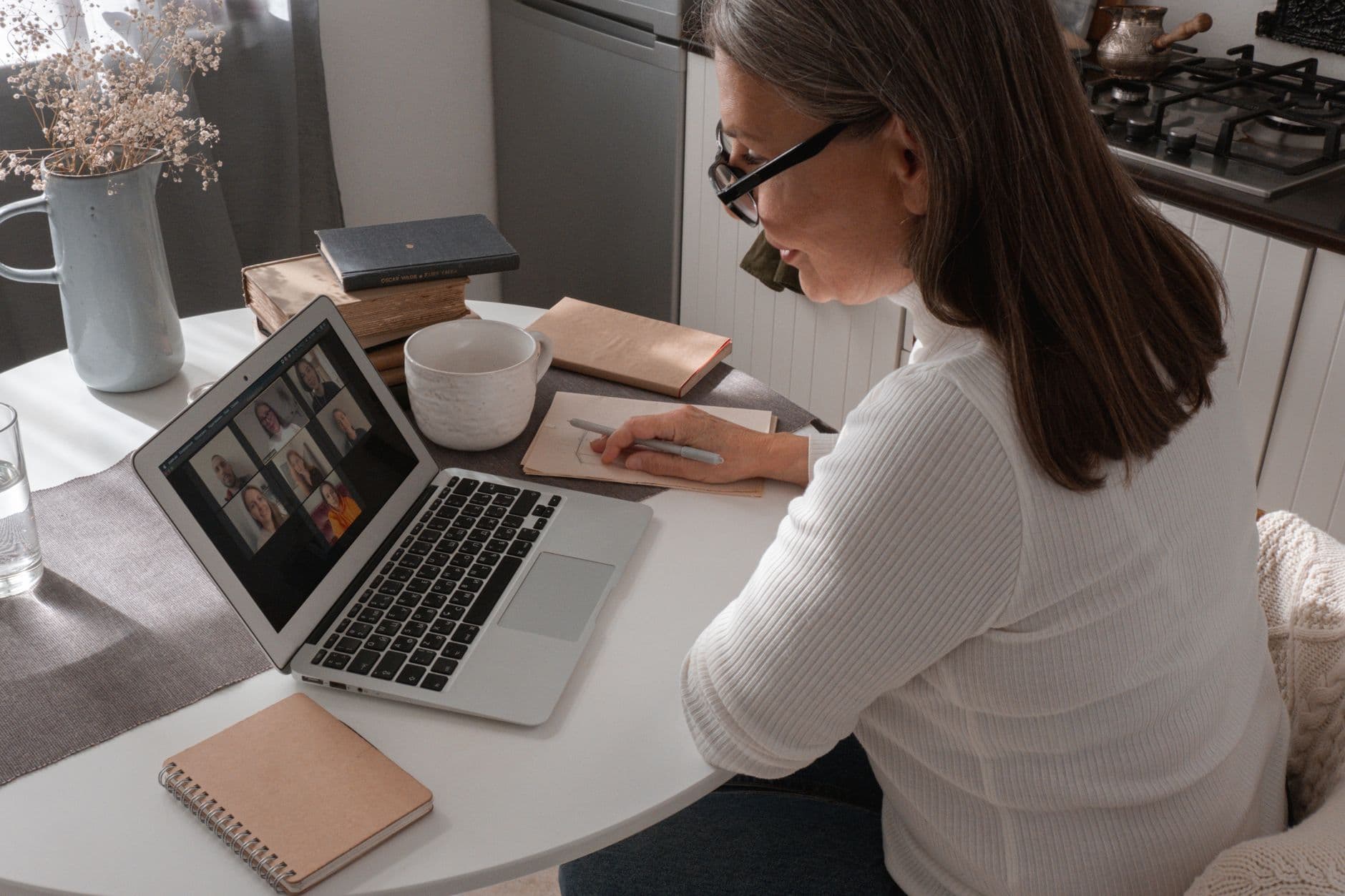 Como se destacar em uma equipe totalmente remota Adult woman participates in a video call, taking notes in a cozy indoor setting.