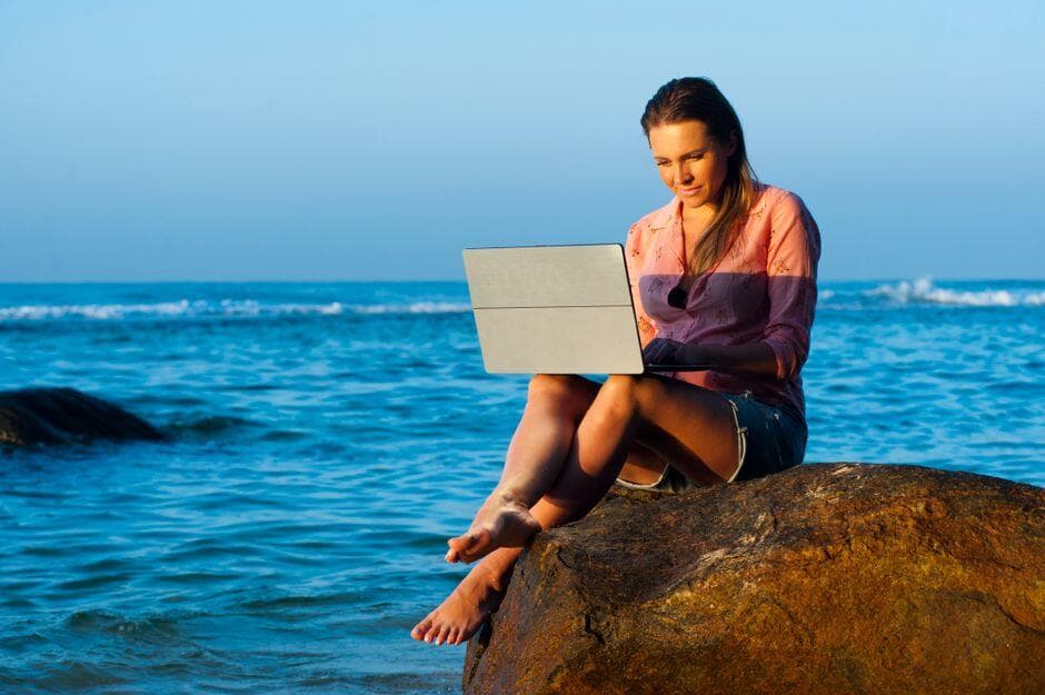 Mulher sentada em uma pedra à beira-mar com notebook no colo