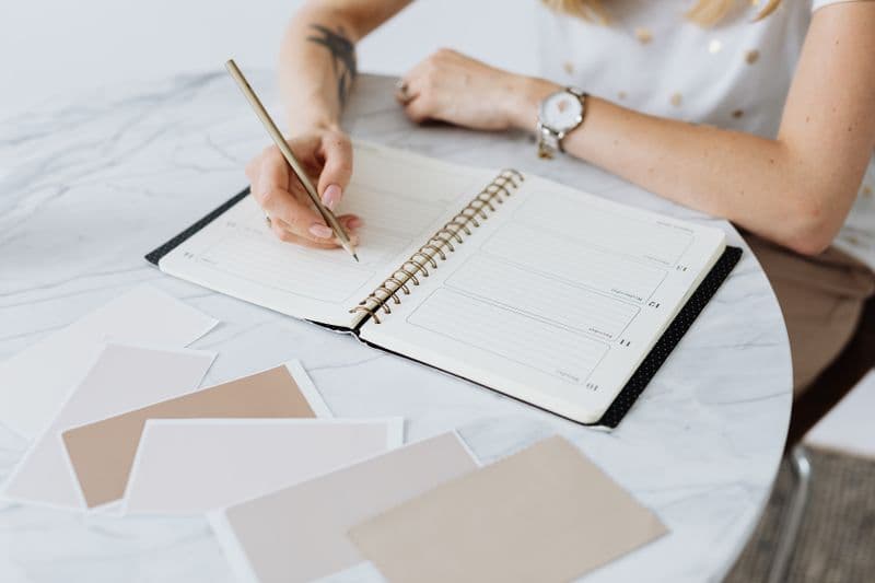 Música ou silêncio para trabalhar: o que funciona melhor A woman writes in a planner on a marble table, surrounded by swatches. Creative and organized workspace.