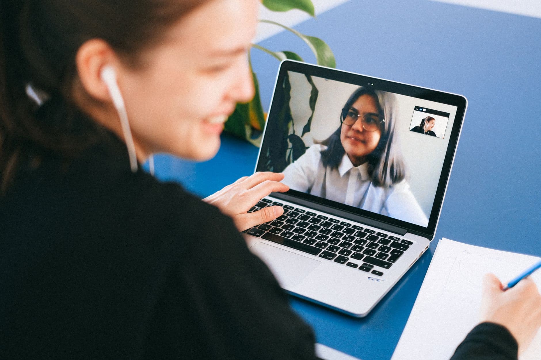 Qual a diferença entre home office e teletrabalho? Woman having a video conference on a laptop, smiling and taking notes.