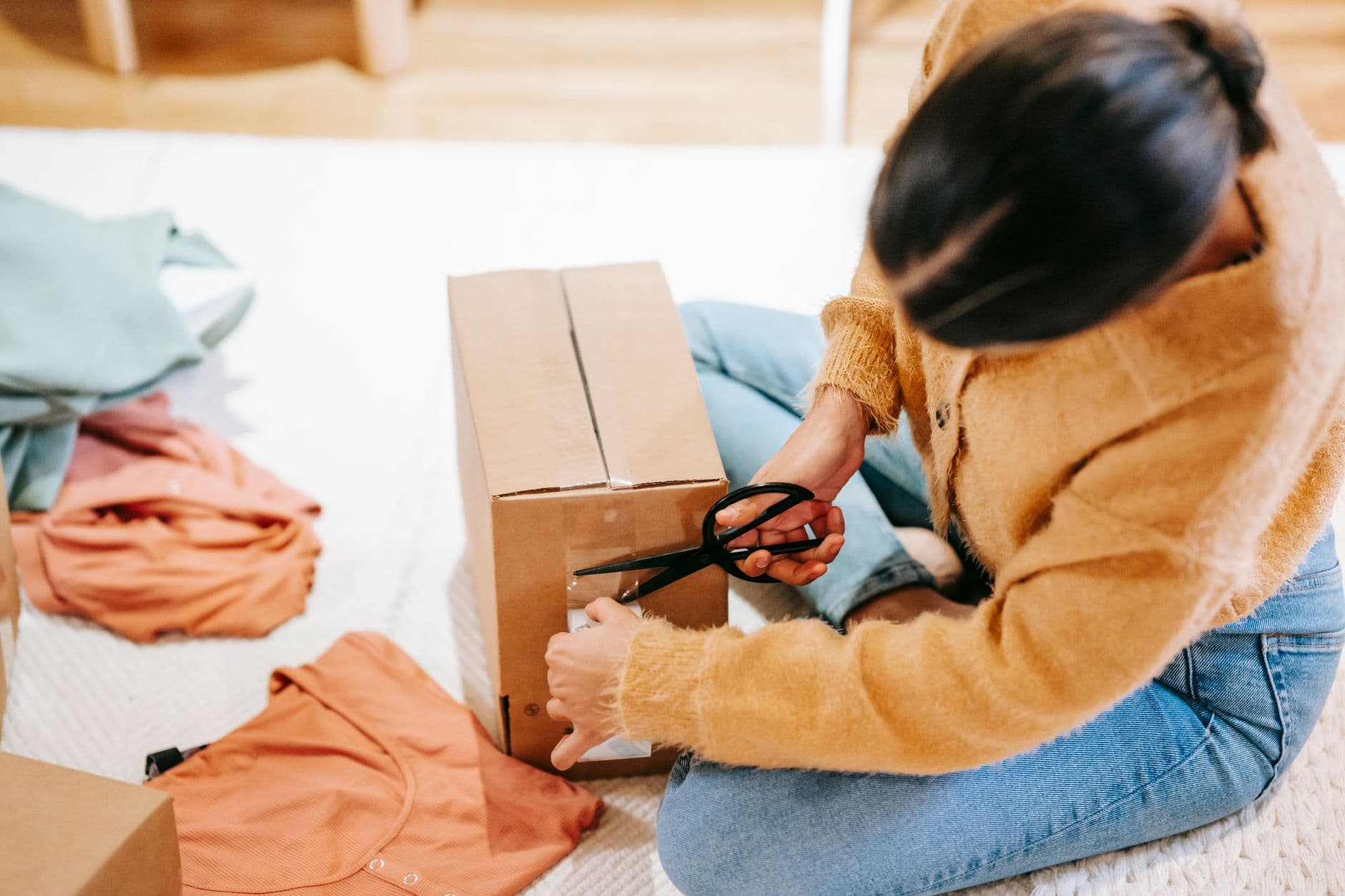 Como empreender trabalhando de casa Crop anonymous female using scissors while packing carton box with parcel on floor among casual clothes