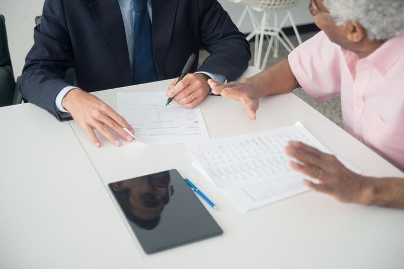 Freelancer: como oferecer pacote mensal ao cliente A business meeting between an elderly client and a consultant discussing documents at an office table.