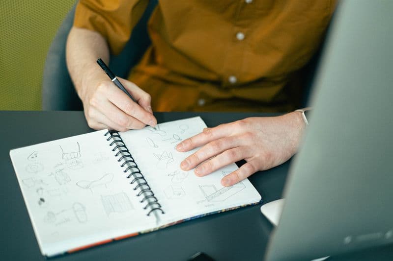 Freelancer de copywriting: portfólio, preços e clientes Close-up of a designer sketching ideas in a spiral notebook at a desk, highlighting creativity.