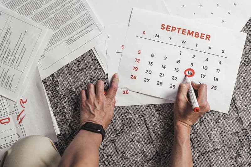 Freelancer: gestão de múltiplos clientes sem perder prazos Hands holding a calendar with September dates encircled, surrounded by documents on a carpet.