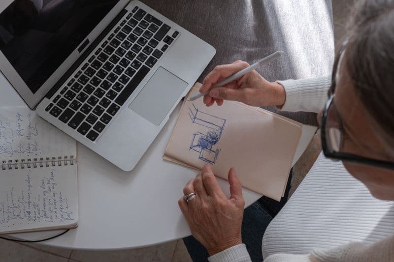 Freelancer ilustrador: como viver de ilustração online An adult woman sketching in a notebook at a desk with her laptop, working from home.