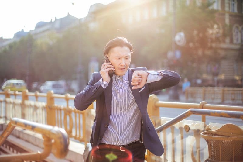 Como lidar com fuso horário em equipe remota internacional Asian businessman in suit checking time while on phone outdoors.