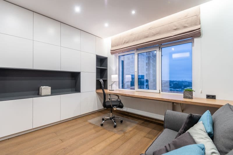 Home office em kitnet: separe trabalho e descanso Armchair near wooden shelf between white cupboards and comfortable sofa with pillow in light stylish room with window at home