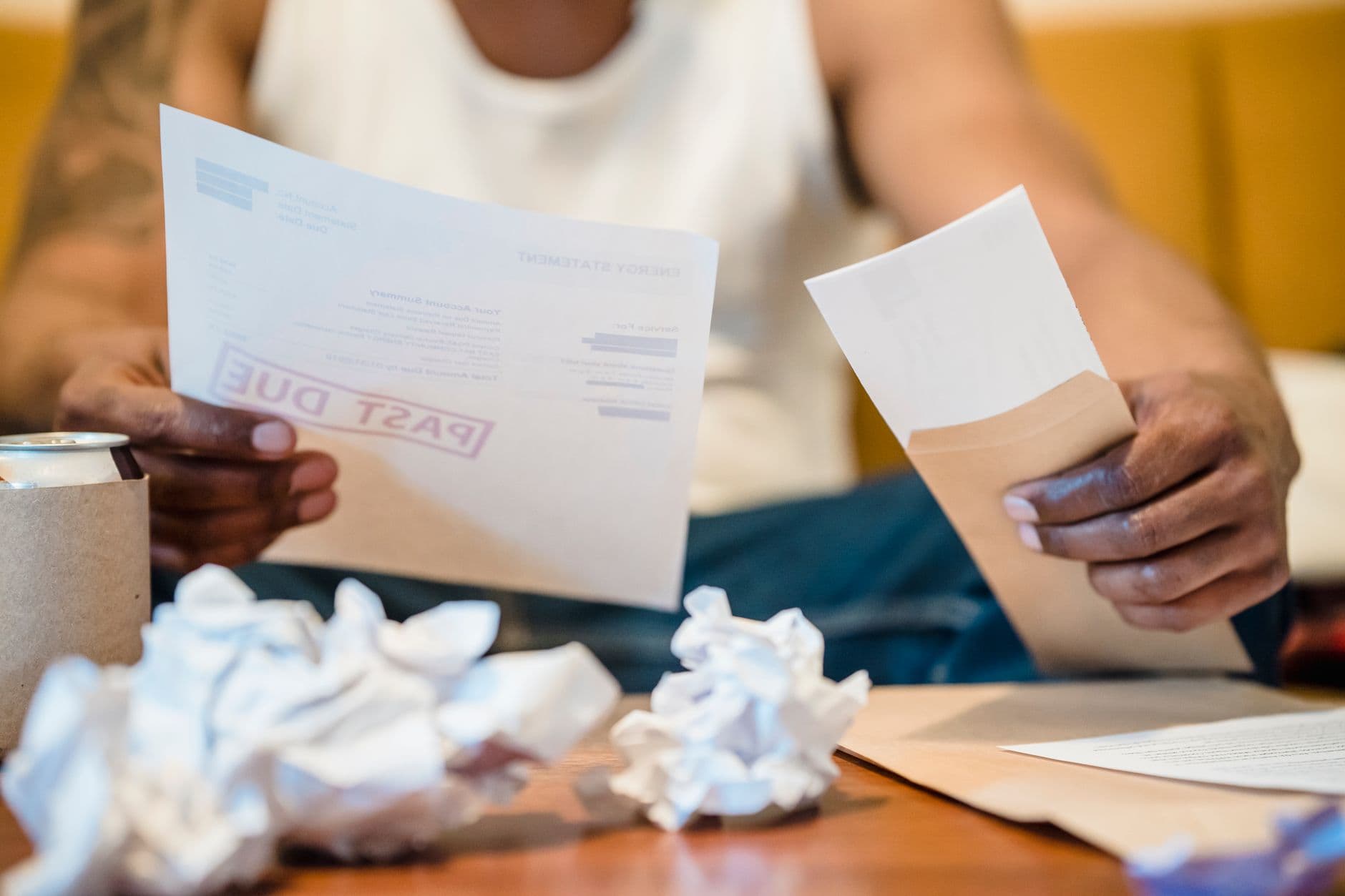 Imposto de renda para quem trabalha remoto para o exterior Man sitting indoors reviewing past due bills with crumpled papers on a coffee table.