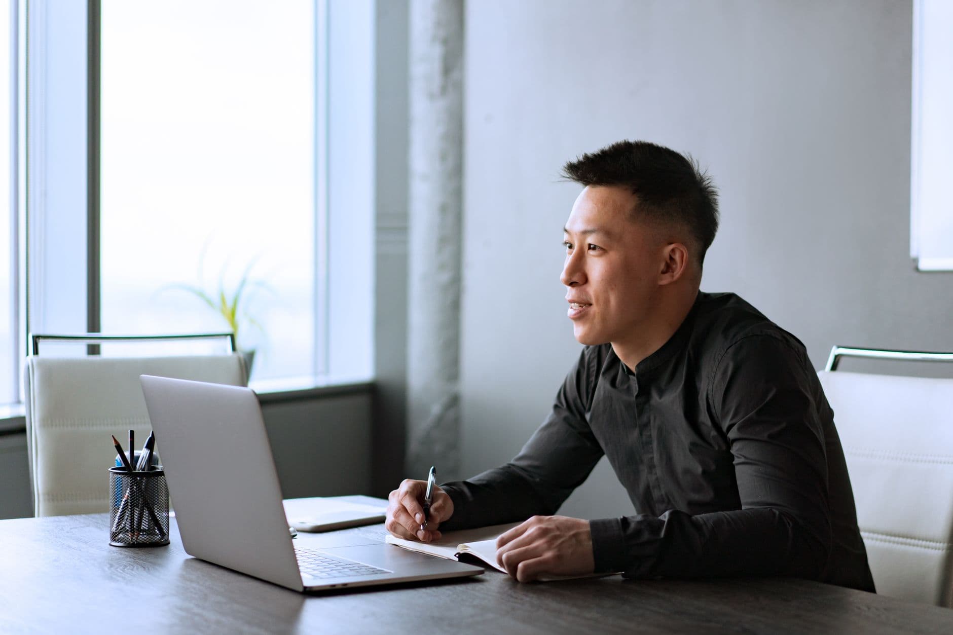 Inglês para trabalho remoto internacional: qual nível? Asian male professional working at a desk in a modern office with a laptop and notepad.