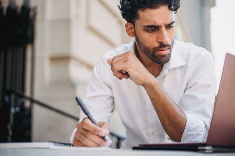 Kanban pessoal: organize o trabalho remoto em colunas simples Focused man in white shirt takes notes while working remotely on his laptop outside, exuding concentration.
