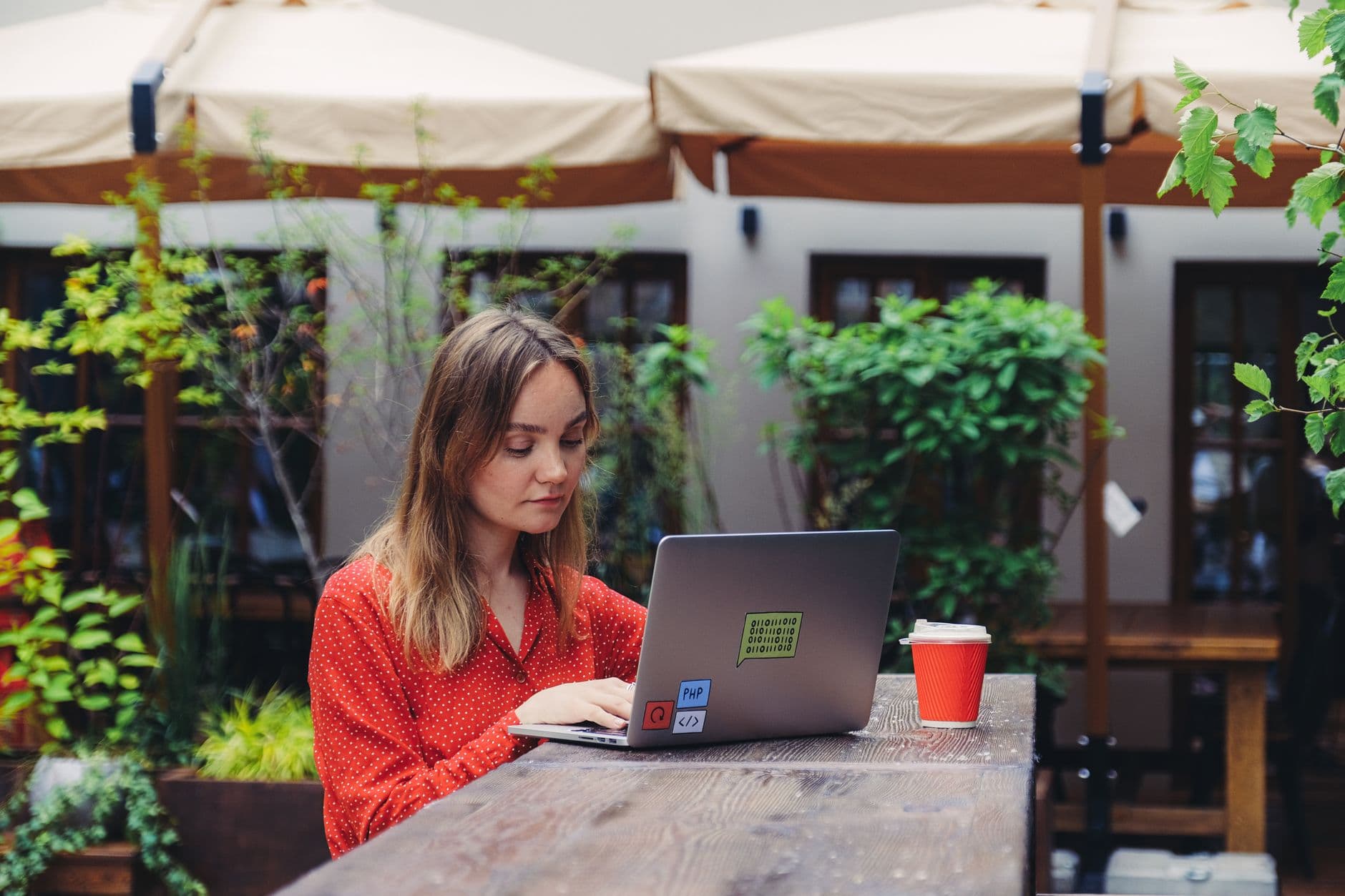 Nômade digital: como começar no Brasil Young woman working on a laptop in an outdoor cafe setting, enjoying a cup of coffee.