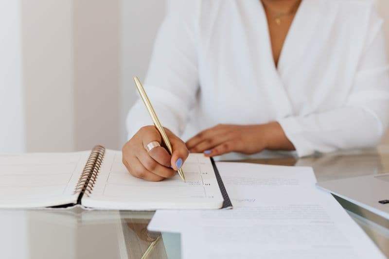 Planner físico ou digital: qual usar no home office Close-up of a person writing notes in a planner with a gold pen, emphasizing organization.