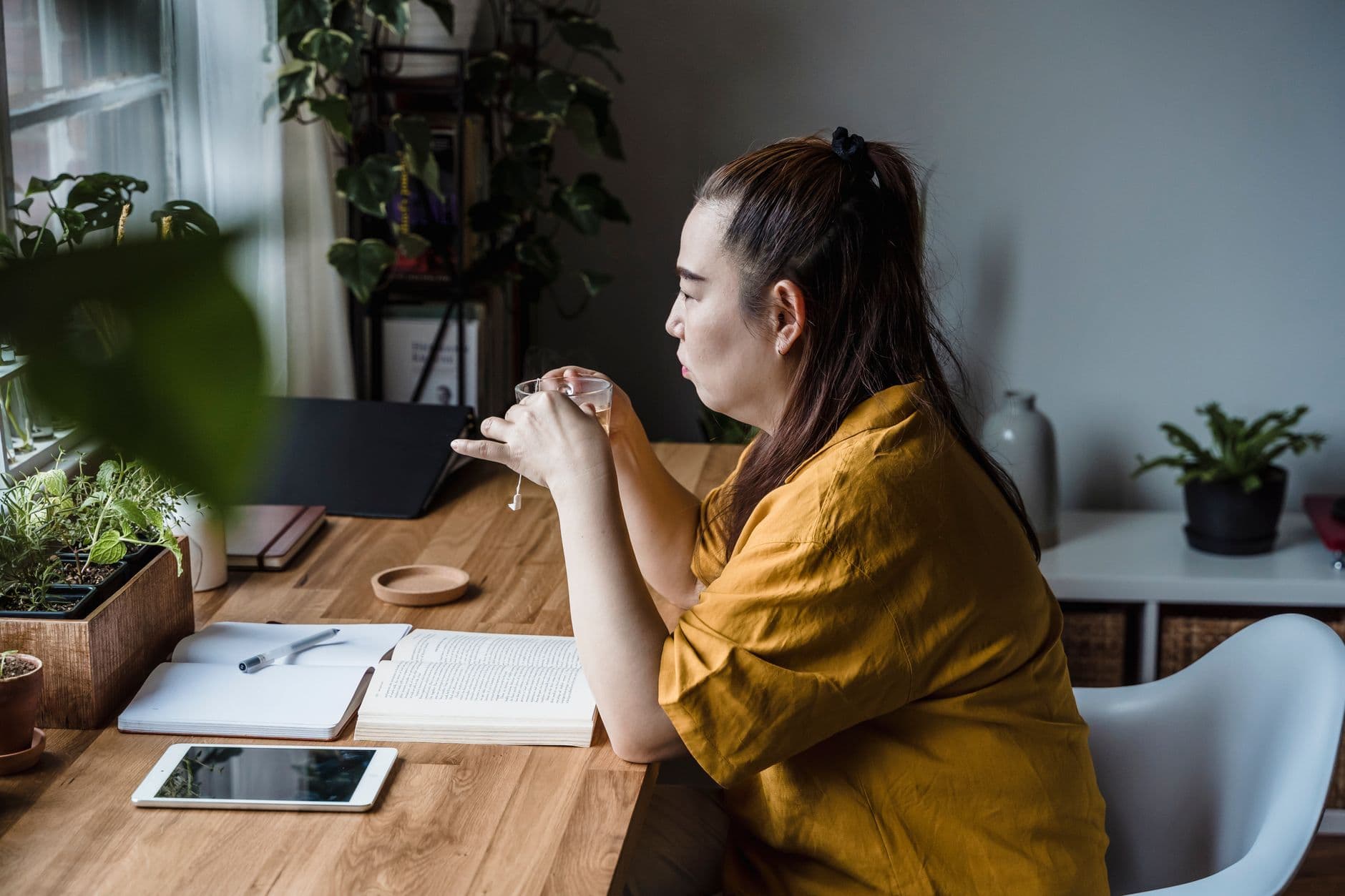 Como manter a produtividade trabalhando em casa Asian woman in yellow blouse enjoying tea while sitting at a wooden desk with plants.