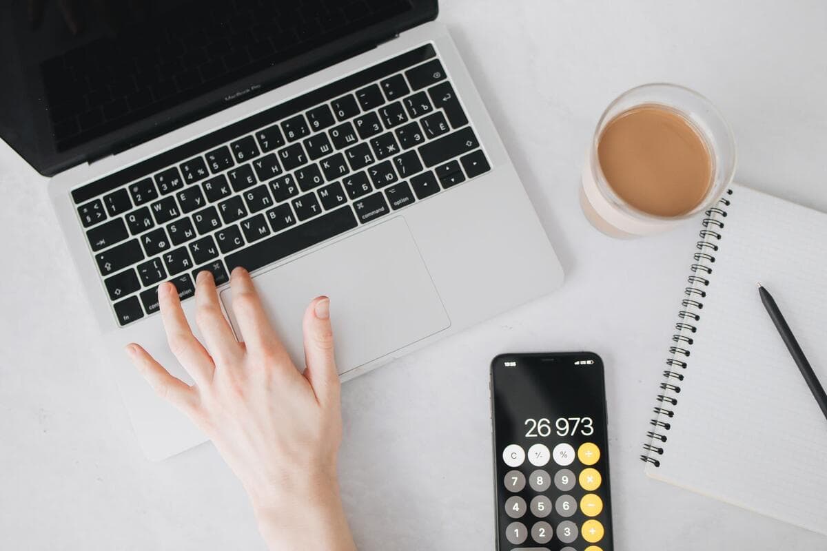Pessoa trabalhando no notebook com calculadora no celular e caderno ao lado