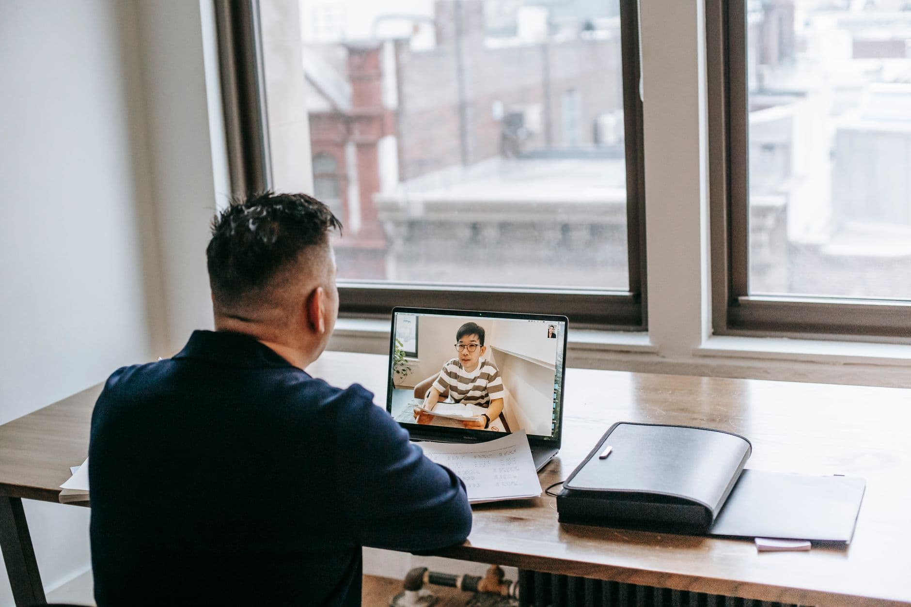Ideias de renda extra trabalhando de casa Back view of male in casual clothes sitting at table on chair while having video chat on laptop with pupil near papers and briefcase in light apartment near window
