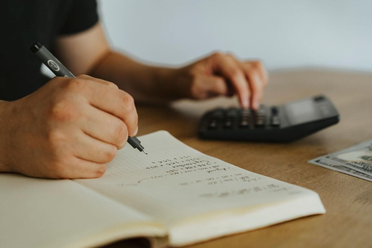 Mãos anotando contas em um caderno ao lado de uma calculadora