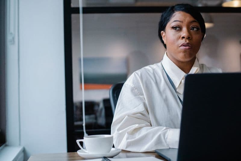 Ruído branco para foco no home office: funciona? Serious African American female employee wearing white formal shirt sitting at working desk with laptop and cup of coffee