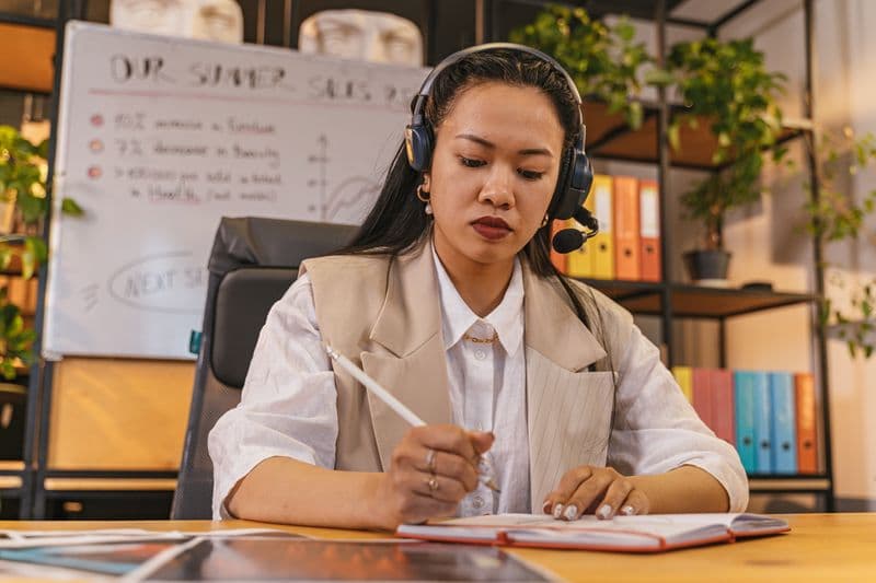 Sedentarismo no trabalho remoto: como se mexer mais Professional woman wearing a headset taking notes in an office setting, focused on work.