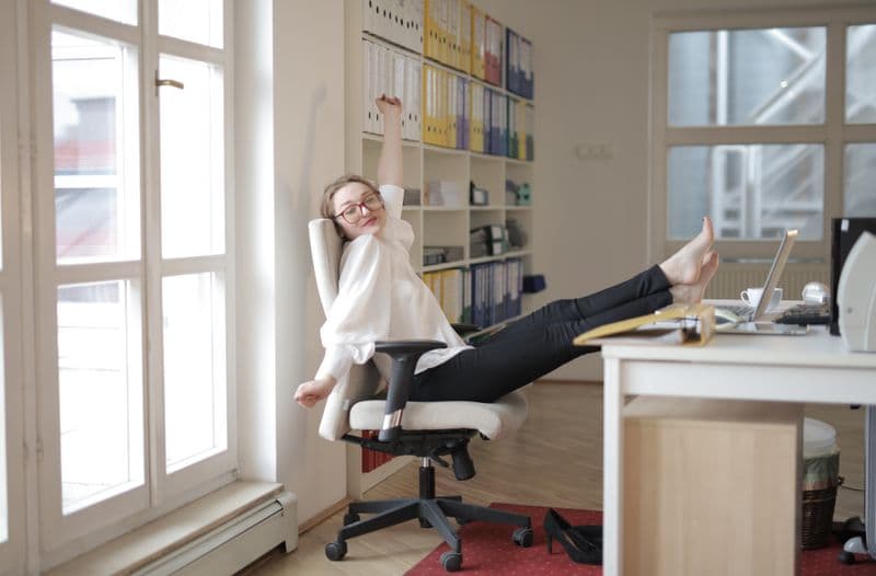 Técnica de ciclos de trabalho e pausa em casa Side view of satisfied young female assistant wearing classy clothes and eyeglasses sitting barefoot on office chair with feet on table and stretching with closed eyes