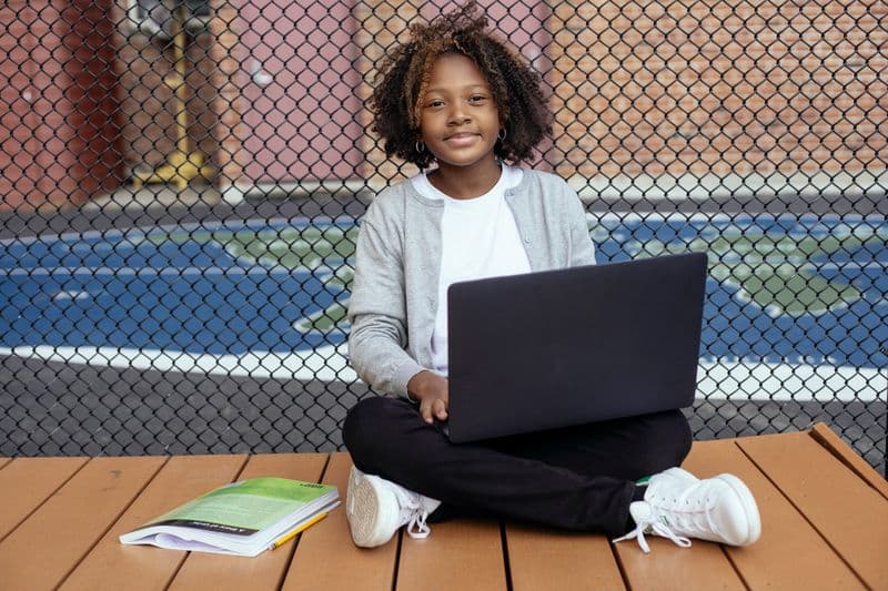 Vagas remotas para professores e pedagogos: onde encontrar Smiling African American schoolchild with Afro hairstyle sitting with netbook on crossed legs while studying and looking at camera near grid fence