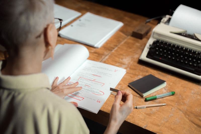 Vagas remotas para jornalistas e produtores de conteúdo An adult woman reviewing a script with red pen marks at a wooden desk with a typewriter.