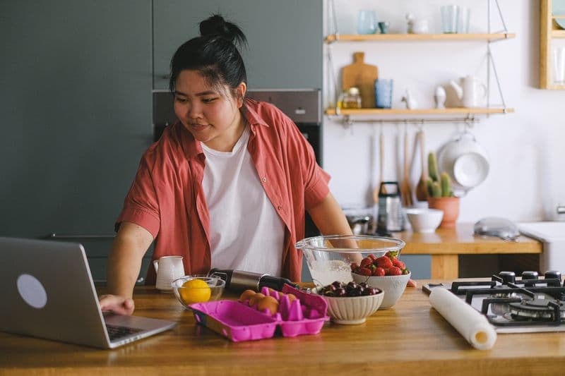 Nutricionista online: como atuar a distância com segurança A young adult using a laptop while preparing food in a modern kitchen setting.