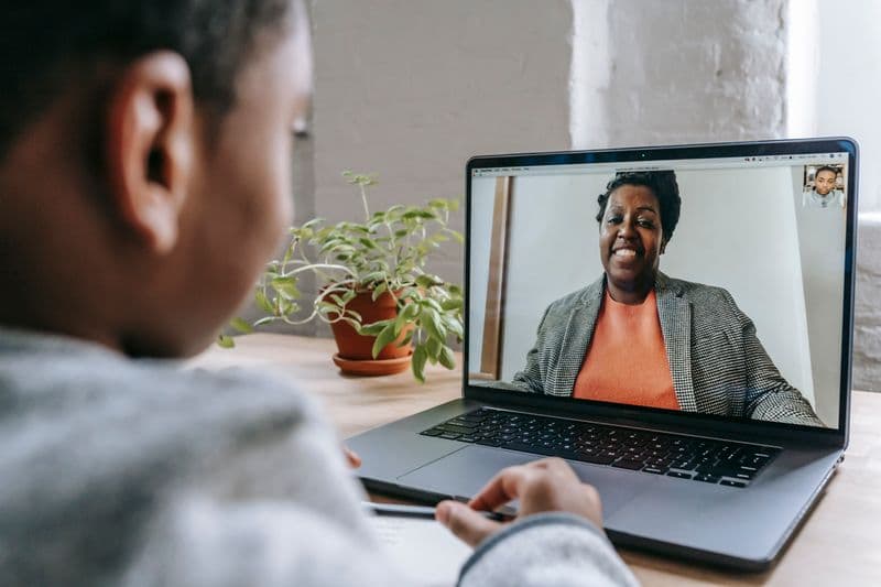 Vagas remotas para quem tem mais de quarenta anos Crop anonymous African American kid sitting at table with modern laptop and listening to female teacher explaining lesson during coronavirus pandemic