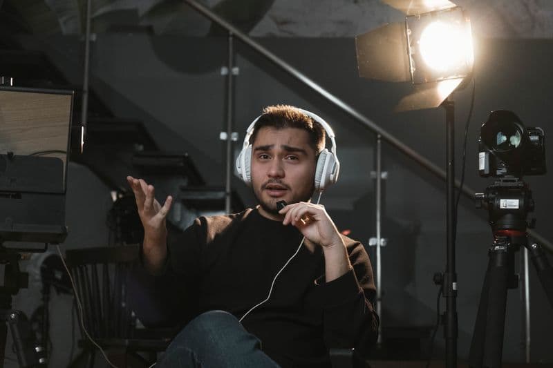 Vagas remotas para recém-formados: como começar Portrait of a man wearing headphones, sitting in a studio, gesturing towards the camera, with a microphone and lighting setup.