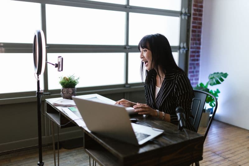 Como escolher uma boa webcam para reuniões Asian woman in office setting recording content with laptop and ring light, showcasing elegant business setup.
