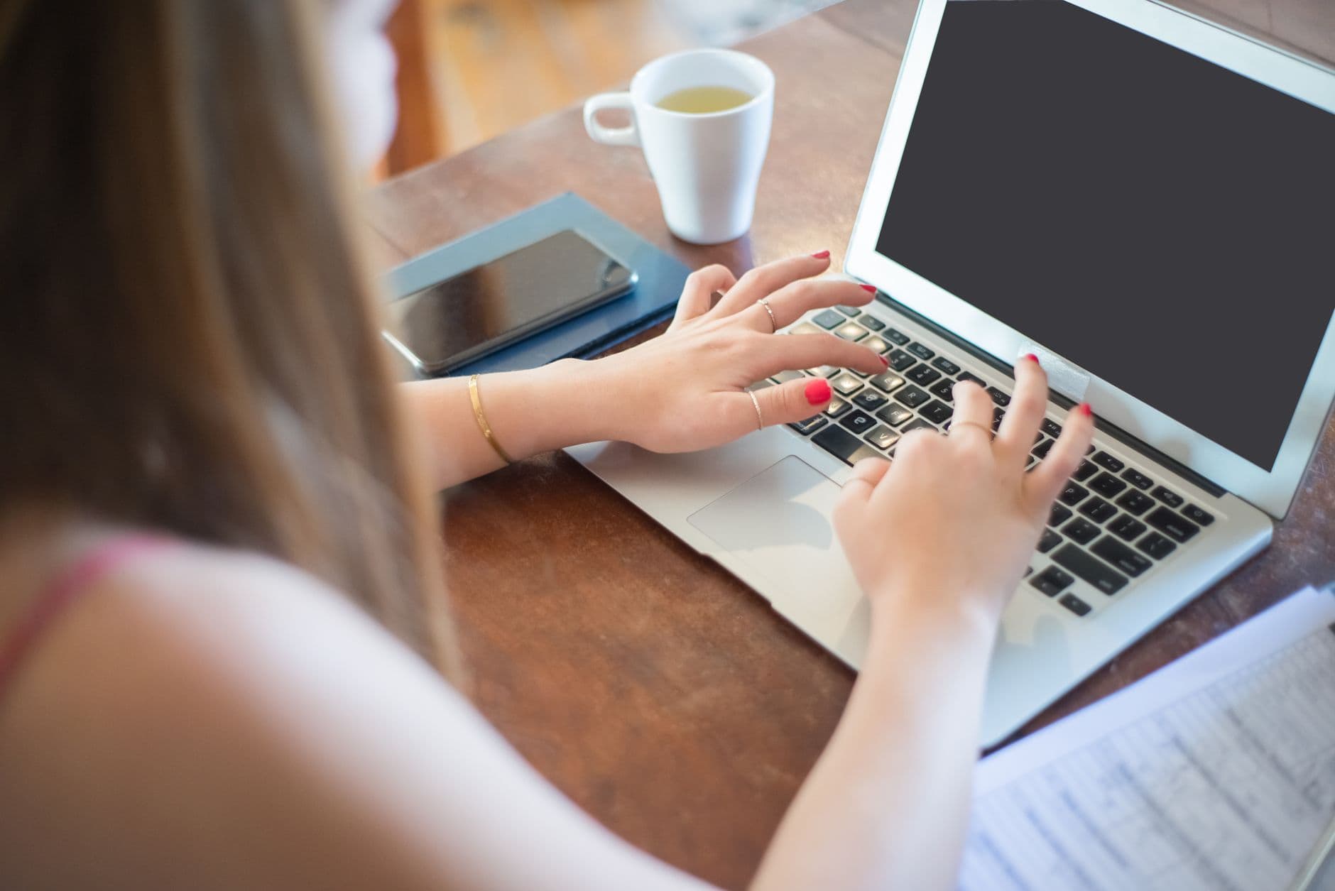 Workana: como começar do zero A woman typing on a laptop with a smartphone and cup on a wooden table. Perfect for remote work themes.